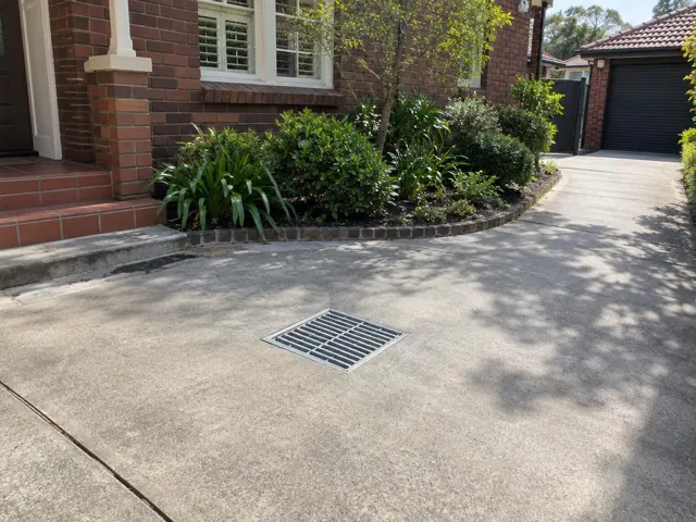 Concrete driveway with a seated stormwater grate beside brick house and garden in Willoughby
